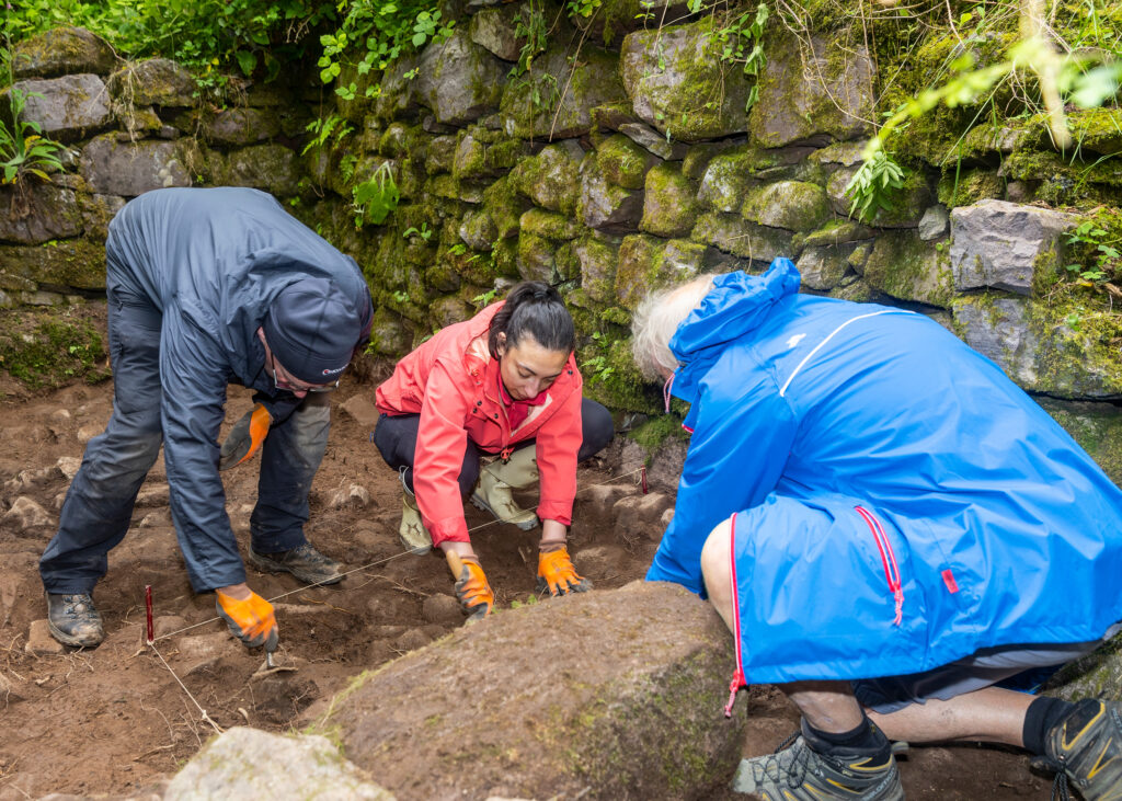 Three archaeologists in raincoats and protective gloves examining the ground at the site of a dig