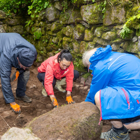 Three archaeologists in raincoats and protective gloves examining the ground at the site of a dig