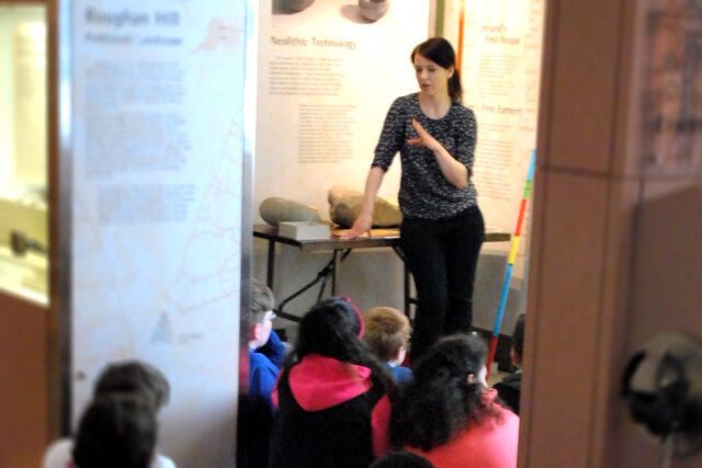 A small group of school children sit facing a teacher in the Museum's long-term exhibition